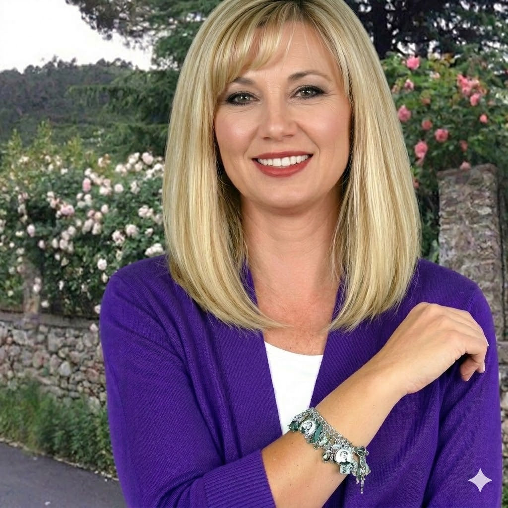 A smiling woman with blonde hair wearing a purple cardigan and a bracelet stands in front of a stone wall with pink flowers.