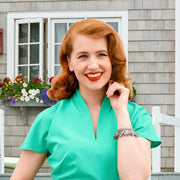 A woman with red hair and a bright smile is posing in front of a house with a flower box. She is wearing a turquoise dress and has a bracelet on her wrist.