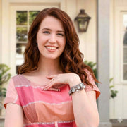 A young woman with long, curly red hair is smiling and posing for a photo in front of a white building with a lantern-style light fixture.