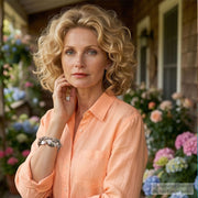 A woman with curly blonde hair wearing a peach-colored blouse stands in front of a house with a flower garden.