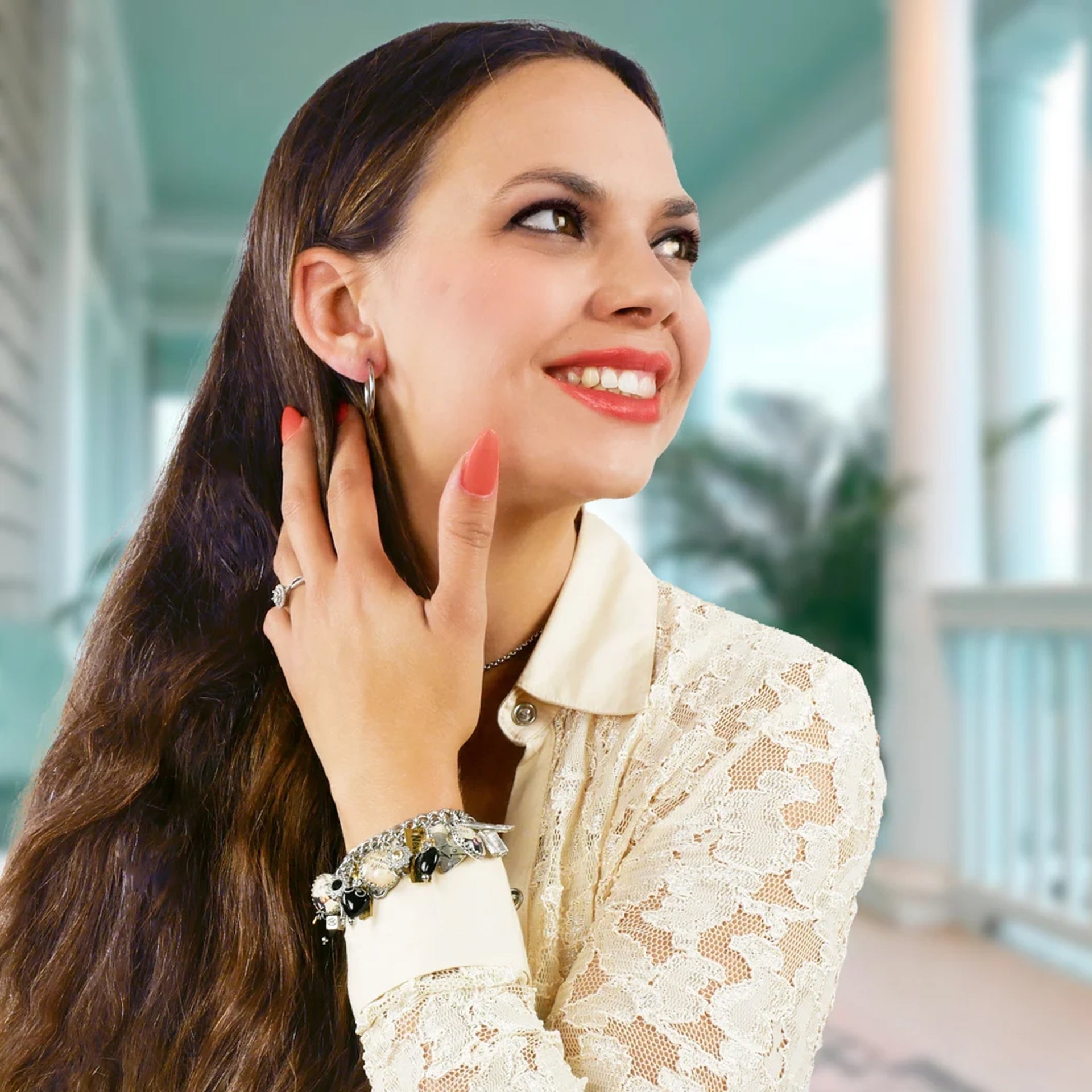A woman with long brown hair is smiling and looking to the side, wearing a white lace blouse and bracelets.