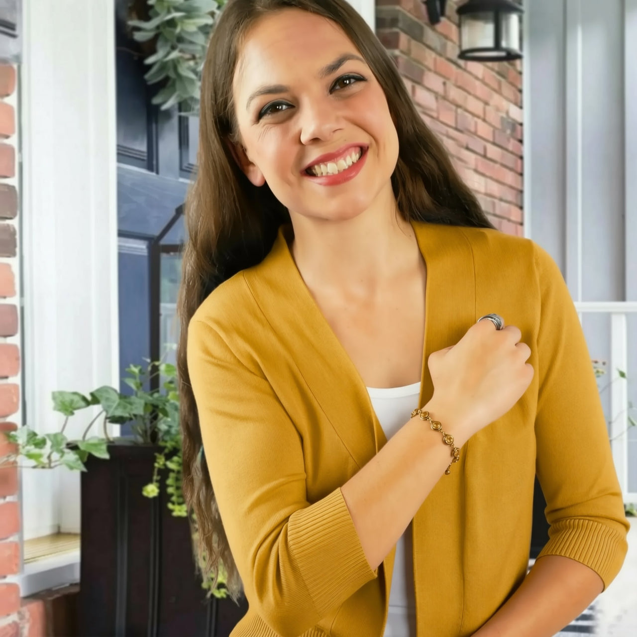 A smiling woman wearing a yellow cardigan stands in front of a brick wall with plants in the background.