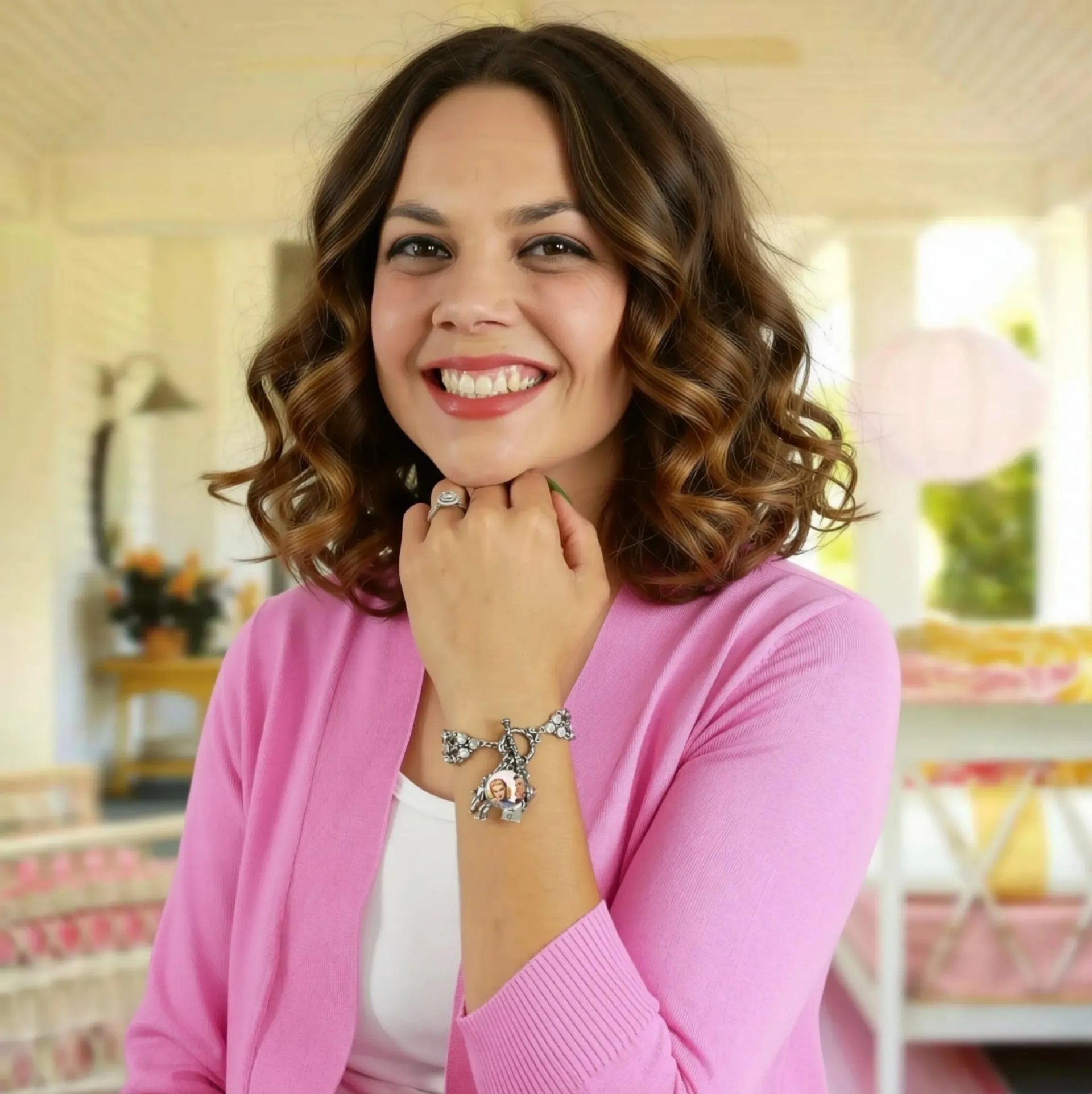 A woman with curly brown hair wearing a pink cardigan and smiling at the camera. She is wearing a bracelet and has a ring on her finger.