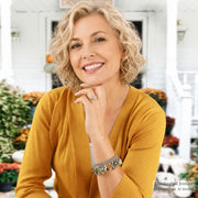 A smiling blonde woman in a mustard yellow cardigan poses in front of a white porch, wearing a silver-toned bracelet and ring. Handcrafted jewelry displayed on an AI model watermark is visible in the bottom right corner.