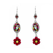 A pair of silver-toned dangle earrings featuring oval Day of the Dead sugar skull medallions and dangling red flower charms with yellow centers, shown on a plain white background.