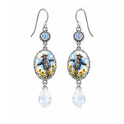 A pair of silver-toned dangle earrings featuring oval charms with an illustrated scarecrow in a blue coat and yellow flowers, with teardrop-shaped clear crystal drops below.