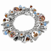 A silver charm bracelet featuring multiple themed charms including a cornucopia, a corn cob with 'Give Thanks' text, a lighthouse charm, a 'Pilgrims' sign, and a '1781' plaque, accented with blue and amber faceted crystals on a white background.