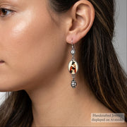A close-up side profile of a woman wearing a dangling earring. The earring has a silver-toned metal setting with a central oval charm featuring a red, white, and black graphic, and a dangling silver bead at the bottom.