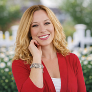 A smiling woman with blonde hair wearing a red cardigan and a bracelet is standing in front of a garden with white flowers.