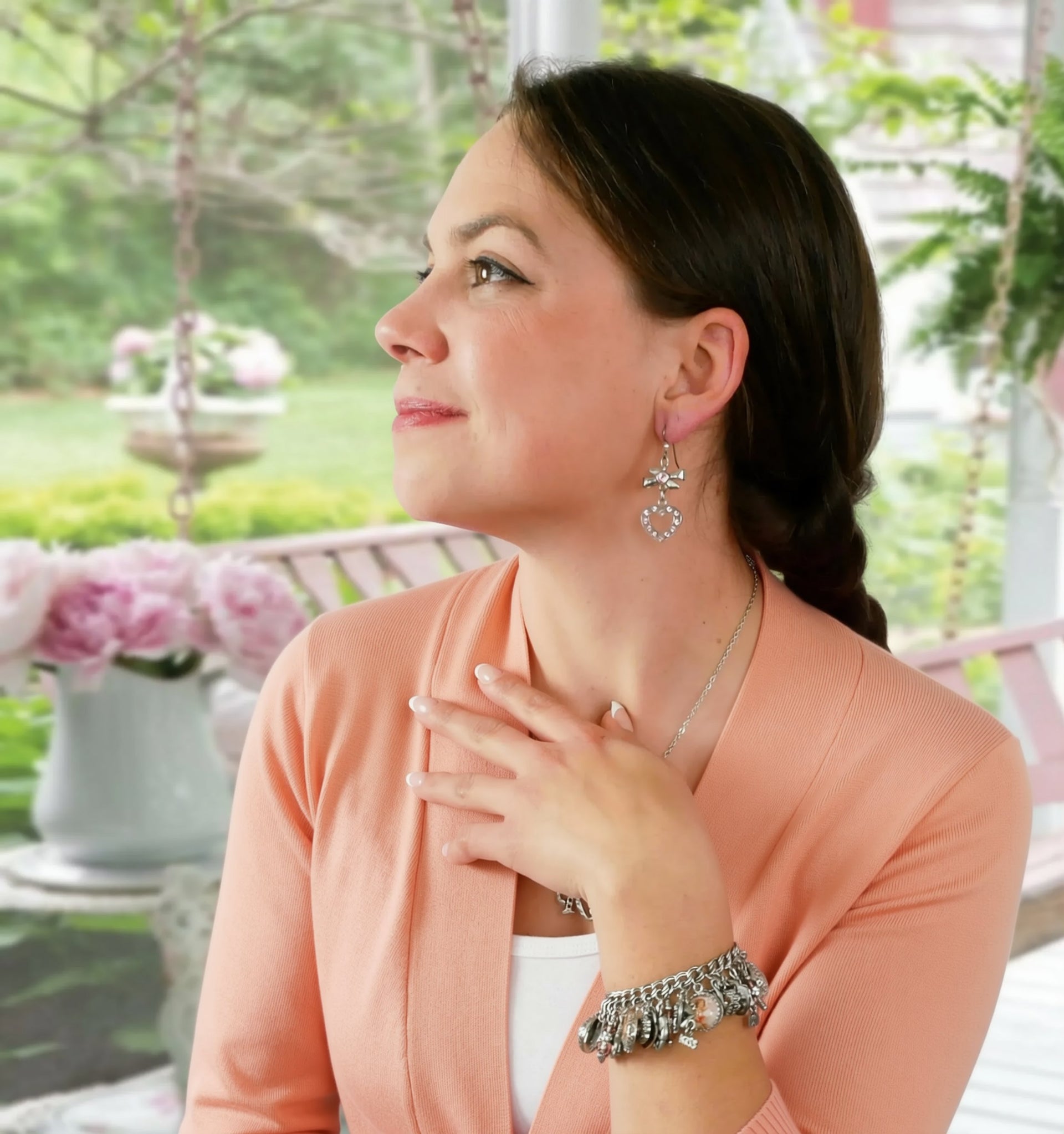 A woman wearing a peach-colored cardigan and jewelry stands in a garden, gazing thoughtfully to the side.