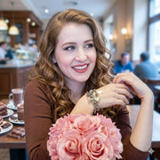 A young woman with curly hair is sitting at a table in a cafe, smiling and holding a bouquet of pink flowers.